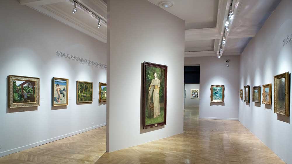 Wnętrze jasnej galerii sztuki z drewnianą parkietową podłogą, sufitowymi reflektorami i ścianami ozdobionymi licznymi oprawionymi obrazami; centralna wolnostojąca ścianka z dużym obrazem przedstawiającym postać w długiej sukni, a po obu stronach korytarza rzędy mniejszych obrazów w ozdobnych ramach.