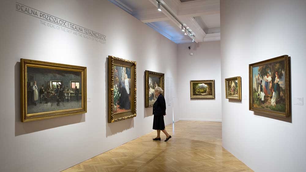 Wnętrze galerii sztuki z jasnymi ścianami i drewnianą podłogą, na których wiszą oprawione obrazy w ozdobnych ramach; w alejce stoi osoba oglądająca ekspozycję, a nad jedną ze ścian widoczny jest napis IDEALNA PRZESZŁOŚĆ I REALNA TERAŹNIEJSZOŚĆ.