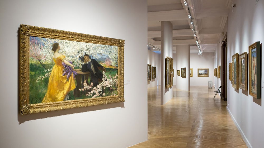 Wnętrze jasnej galerii sztuki z drewnianą podłogą i sufitowymi reflektorami, długim korytarzem i białymi ścianami z wieloma obrazami — po lewej duży obraz w ozdobnej złotej ramie przedstawiający kobietę w żółtej sukni i postać w ciemnym stroju, po prawej oraz w głębi kolejne oprawione płótna i filary.