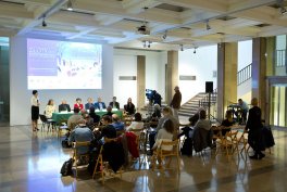 Duża jasna sala z ekranem projekcyjnym na ścianie, przed którym siedzi kilkuosobowy panel przy długim stole, publiczność na składanych krzesłach ustawionych w półokręgu, operator kamery i schody po prawej stronie.