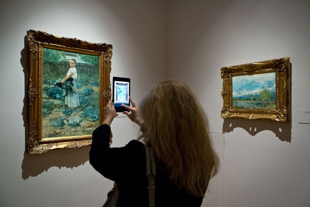 Osoba widoczna od tyłu o długich jasnych włosach robi smartfonem zdjęcie dwóm złoconym, ozdobnym ramom obrazów na ścianie galerii; po lewej widoczny obraz przedstawia dziewczynkę stojącą w ogródku z kapustą, po prawej mniejszy pejzaż z górami i wodą.