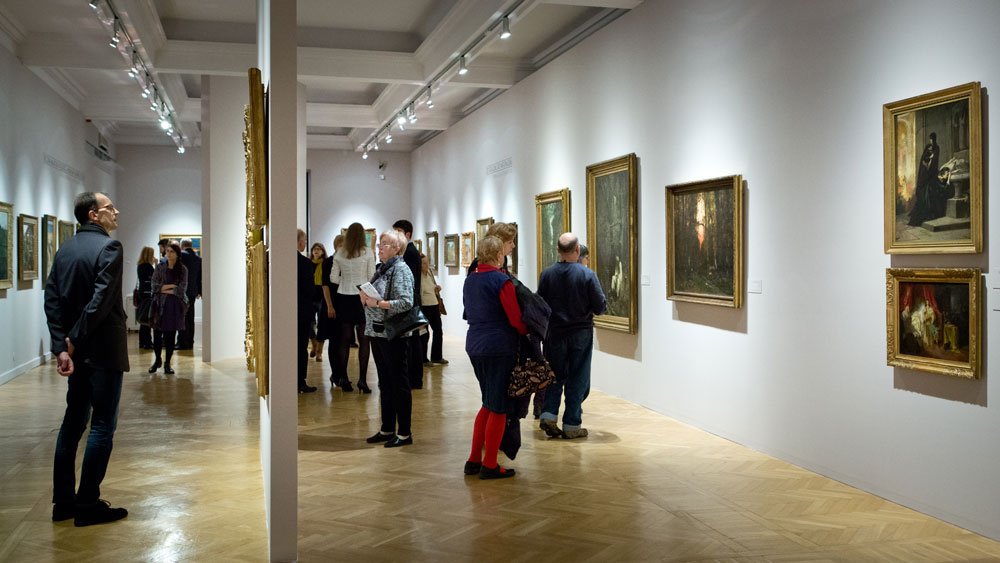 Galeria sztuki o białych ścianach z obrazami w ozdobnych ramach, wąską ścianą dzielącą przestrzeń, kilkoma zwiedzającymi oglądającymi dzieła, sufitowymi reflektorami i jasną jodełkową podłogą.