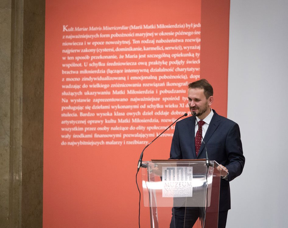 Mężczyzna w granatowym garniturze i bordowym krawacie stoi przy przezroczystej mównicy z mikrofonami i widocznym logo muzeum, na tle dużego czerwonego panelu z białym tekstem oraz jasnej ściany, z marmurową kolumną po lewej.