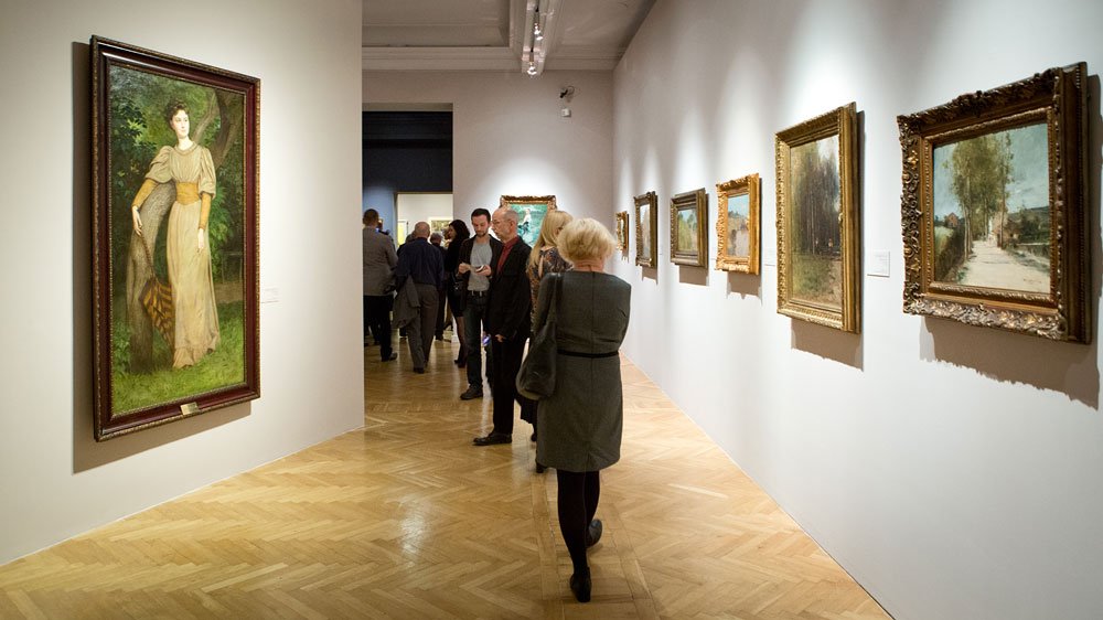 Wnętrze galerii sztuki z kilkoma osobami oglądającymi obrazy zawieszone na białych ścianach; po lewej duży portret kobiety, po prawej rząd mniejszych pejzażowych obrazów w ozdobnych ramach i jasna drewniana podłoga.