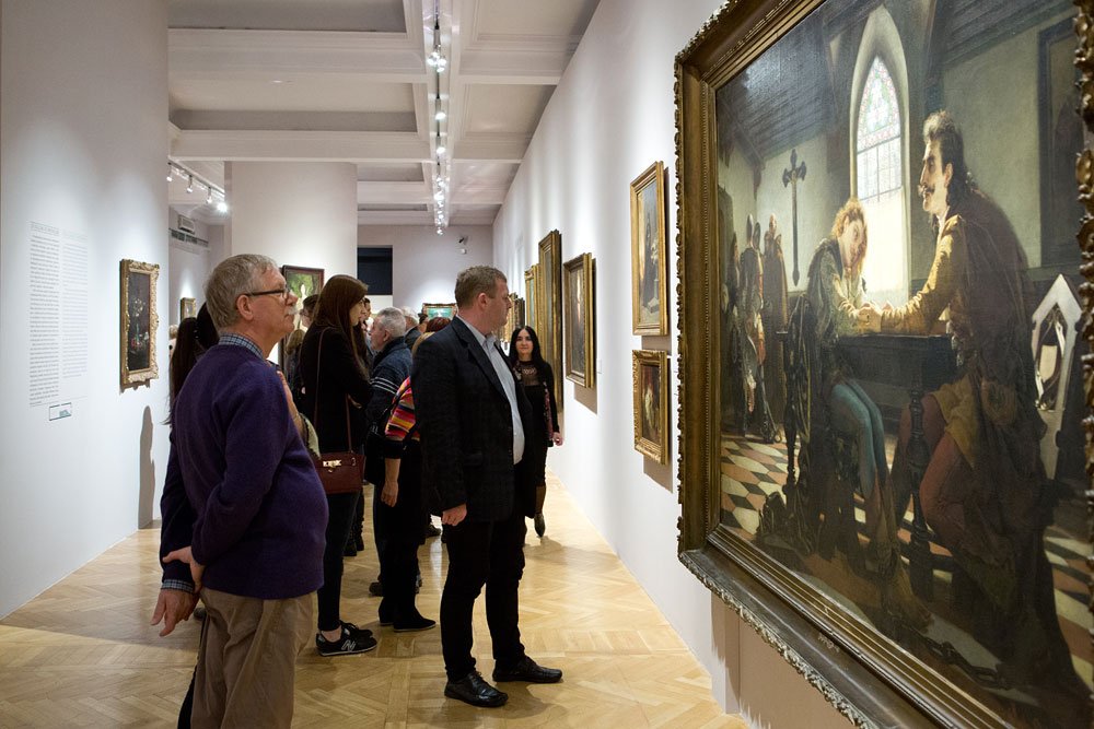 Grupa osób stoi i ogląda obrazy w długim białym wnętrzu galerii z drewnianą chevronową podłogą i sufitowym oświetleniem, po prawej duże, zdobione ramy z historycznym obrazem przedstawiającym mężczyzn siedzących przy stole.