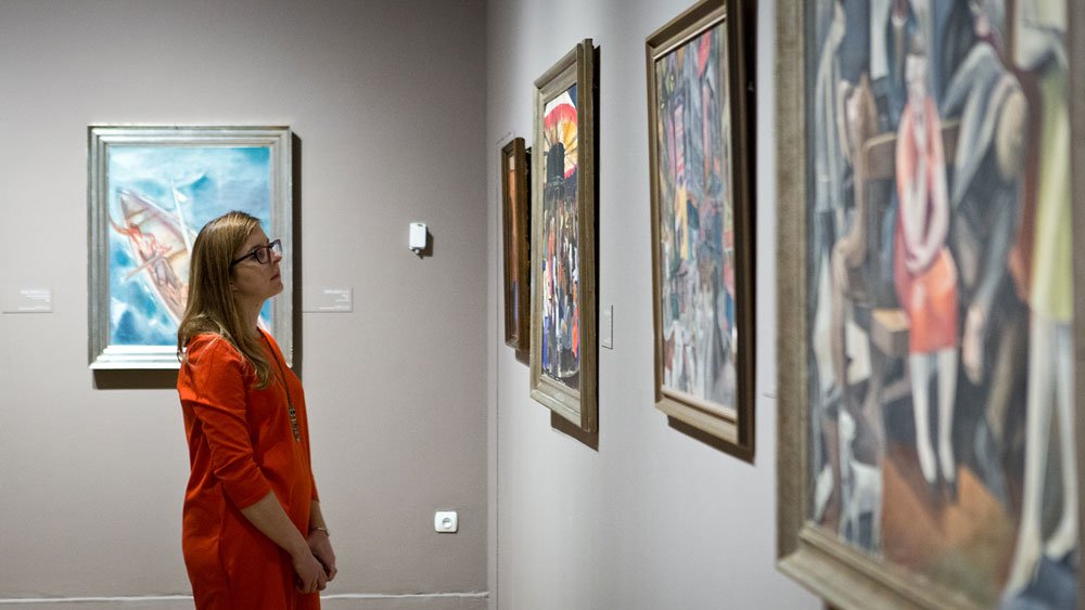 Kobieta w pomarańczowej sukience i okularach stoi z profilu w galerii sztuki przed jasnoszarą ścianą z kilkoma oprawionymi obrazami w drewnianych ramach.