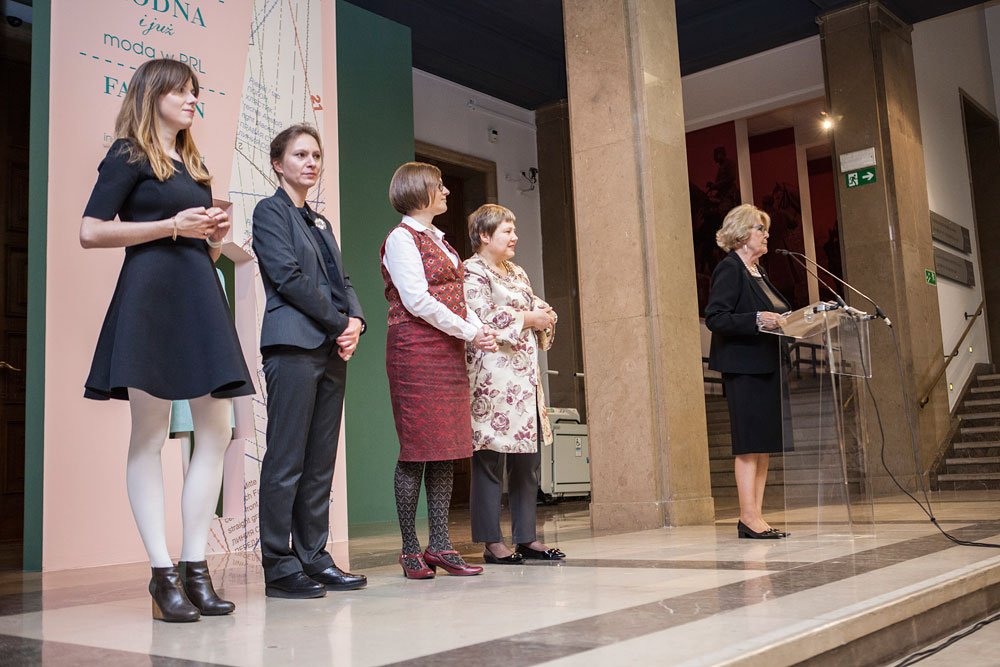 Pięć kobiet stojących na scenie w reprezentacyjnym hallu: cztery ustawione obok siebie w różnych strojach (czarna sukienka z białymi rajstopami, ciemny garnitur, czerwona sukienka z białą koszulą, jasna tunika w kwiaty z ciemnymi spodniami) oraz kobieta przy przezroczystym pulpicie z mikrofonem, w tle kolumny, marmurowa posadzka, baner z fragmentem tekstu i schody.