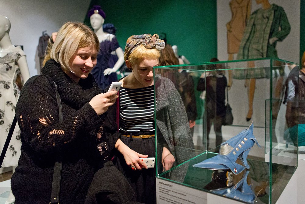 Dwie kobiety stojące przy szklanej gablocie w muzeum: po lewej blondynka w czarnym ażurowym swetrze uśmiecha się i trzyma telefon, po prawej krótko obcięte włosy z apaszką, w pasiastym topie i szarym płaszczu także trzyma telefon i ogląda w gablocie niebieskie awangardowe buty, w tle manekiny i ilustracje ubrań na zielonej ścianie.