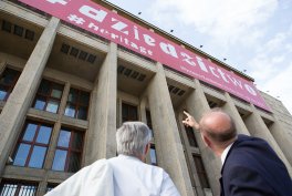 Dwóch mężczyzn widocznych od tyłu — jeden z siwymi włosami, drugi łysy wskazujący palcem — stoi przed dużym kamiennym budynkiem z wysokimi kolumnami, rzędami okien i różowym banerem z białym napisem na elewacji pod pochmurnym niebem.