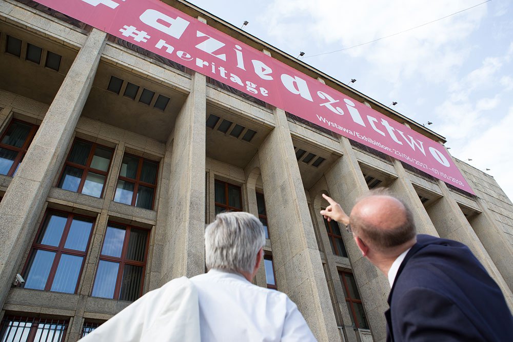 Zdjęcie z niskiego kąta pokazujące fasadę budynku z wysokimi betonowymi kolumnami i dużym różowym banerem z napisem "#heritage", z dwoma mężczyznami widzianymi od tyłu — jeden w białej koszuli, drugi w ciemnej marynarce wskazujący w górę.