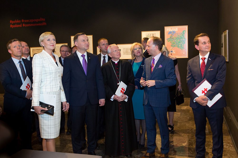 Grupa osób w formalnych strojach stoi w galerii sztuki przed ciemną ścianą z oprawionymi grafikami; w centrum kobieta w białym kostiumie trzyma czarną kopertówkę, obok mężczyzna w granatowym garniturze i mężczyzna w czarnym stroju z czerwonymi wstawkami, kilku innych mężczyzn w garniturach trzyma broszury z czerwonym znakiem.