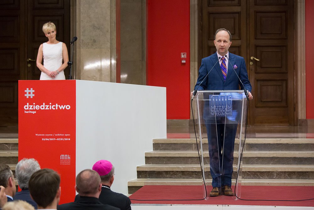 Ubrany w granatowy garnitur mężczyzna stoi za przezroczystą mównicą z mikrofonami i napisem „MUZEUM”, po lewej kobieta w białej sukience przy dużym czerwono‑białym panelu z napisem „dziedzictwo heritage”, w tle marmurowe schody i drewniane drzwi, a na pierwszym planie widać tyły głów widowni, w tym osobę z różowym nakryciem głowy.