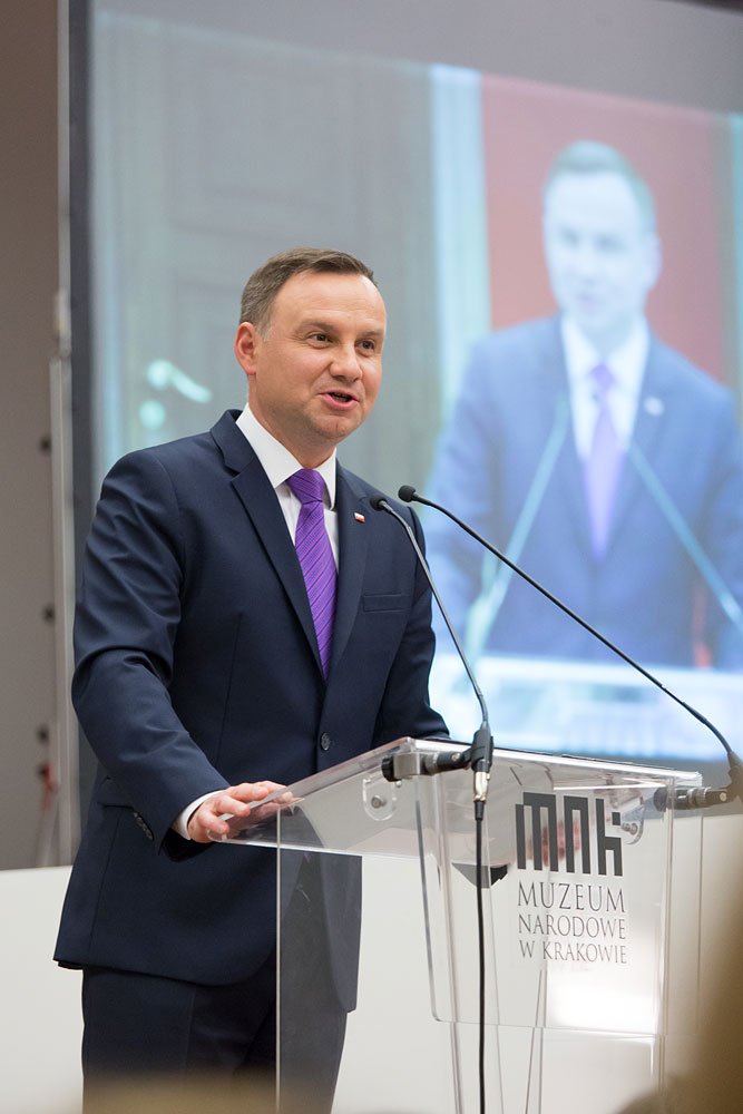 Mężczyzna w granatowym garniturze i fioletowym krawacie przemawia przy przezroczystej mównicy z dwoma mikrofonami, na której widoczne jest logo i napis Muzeum Narodowe w Krakowie, a za nim ekran z rozmytą projekcją jego postaci.