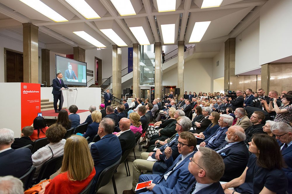 Mówca stojący za przezroczystą mównicą obok czerwonego panelu z napisem "dziedzictwo", przemawiający do dużej publiczności siedzącej w rzędach w jasnym holu z kolumnami, panelami świetlnymi w suficie, schodami i ekranem projekcyjnym pokazującym mówcę, przy prawym boku sali fotografowie i kamery.