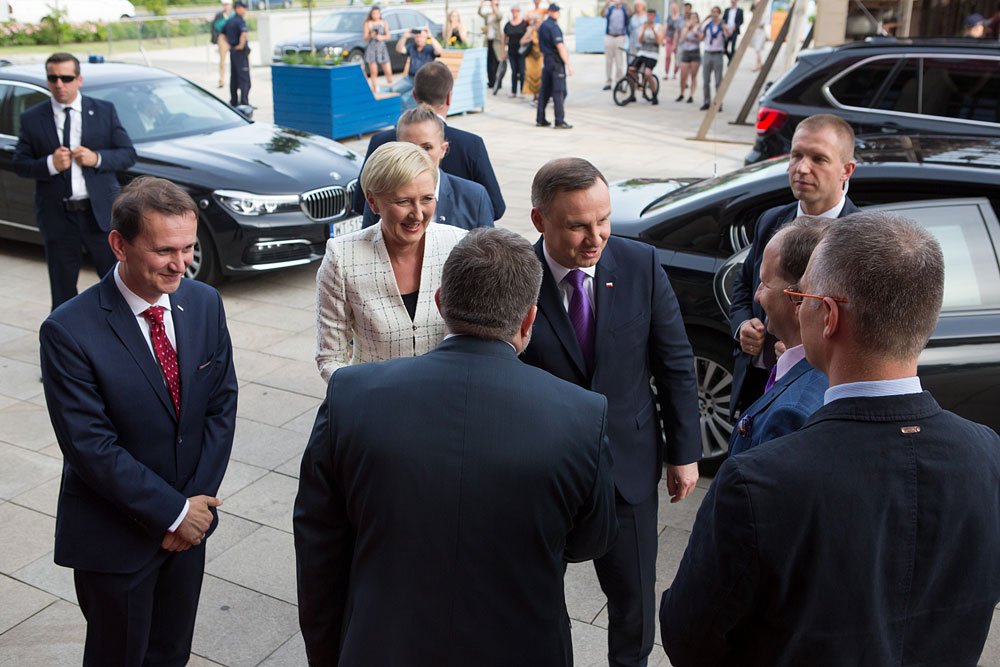 Kilka formalnie ubranych osób — mężczyzn w garniturach i kobieta w jasnej marynarce — stoi i rozmawia na kamiennej nawierzchni obok zaparkowanych czarnych samochodów, w tle przechodnie i ochroniarze.