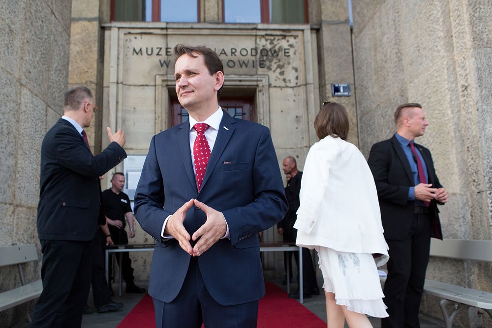 Mężczyzna w granatowym garniturze i czerwonym krawacie z białymi kropkami stoi na czerwonym dywanie przed kamiennym wejściem z widocznym napisem "MUZEUM NARODOWE", z dłońmi złączonymi w kształt trójkąta, a wokół niego kilka osób w formalnych strojach rozmawia i przechodzi obok.