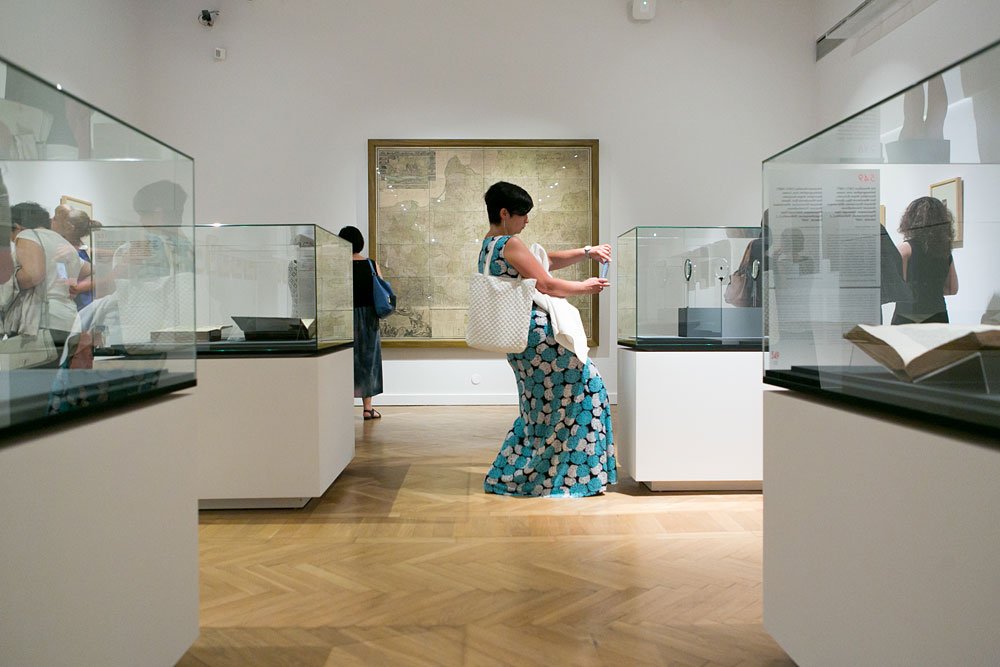 Wnętrze galerii muzealnej z jasnym parkietem, przeszklonymi gablotami na białych postumentach i dużą oprawioną mapą na ścianie; pośrodku kobieta w długiej niebiesko-białej sukni w kwiaty z dużą białą torbą trzyma poziomo smartfon nad gablotą, w tle inni zwiedzający i otwarta książka w gablocie na pierwszym planie po prawej.