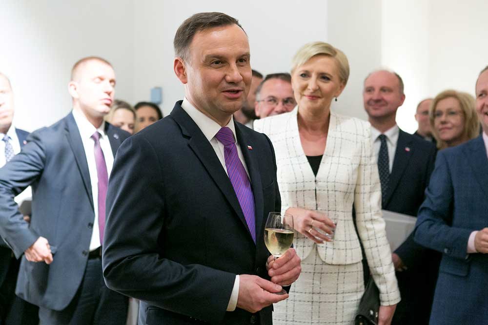 Mężczyzna w ciemnym garniturze i fioletowym krawacie stoi na pierwszym planie trzymając kieliszek białego wina, za nim kobieta w białym żakiecie w kratę z szklanką w ręku, a w tle grupa osób w formalnych strojach w jasnym wnętrzu.