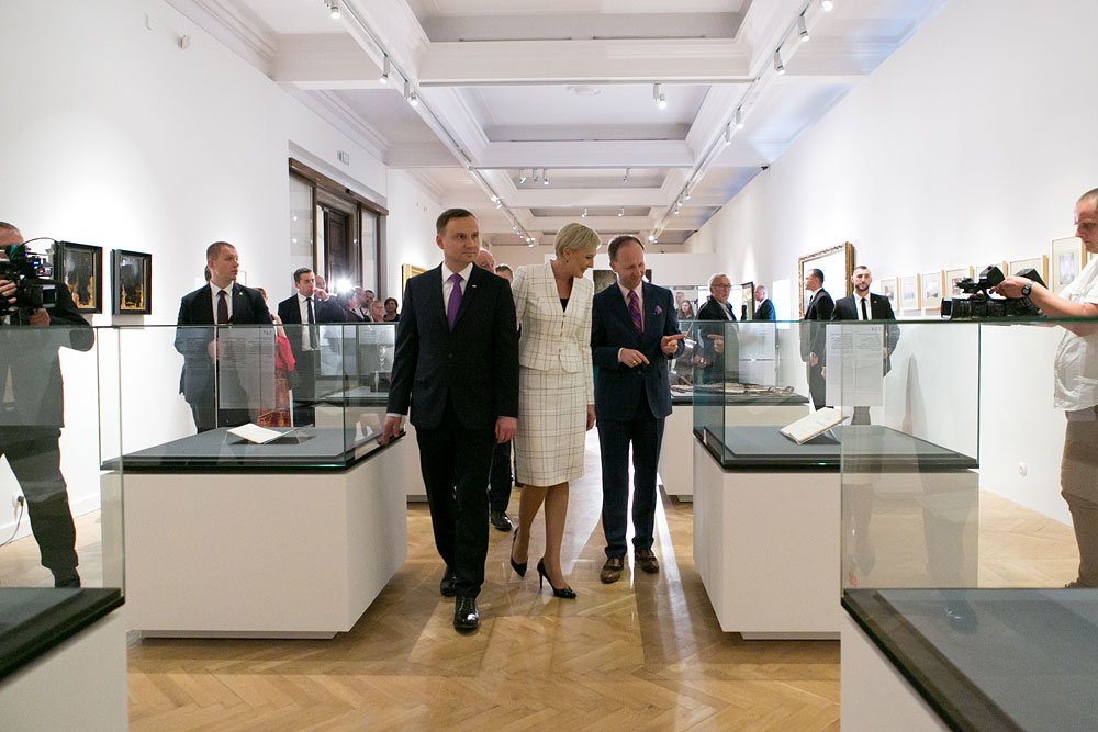Wnętrze galerii z drewnianą jodełkową podłogą i sufitowymi reflektorami, rząd przeszklonych gablot z dokumentami i książkami, przez środek idą trzy formalnie ubrane osoby (mężczyzna w ciemnym garniturze i fioletowym krawacie po lewej, kobieta w jasnym kraciastym kostiumie w środku, mężczyzna w ciemnym garniturze i fioletowym krawacie po prawej), a po bokach stoją ochroniarze, kamerzyści i zwiedzający; na białych ścianach oprawione obrazy.