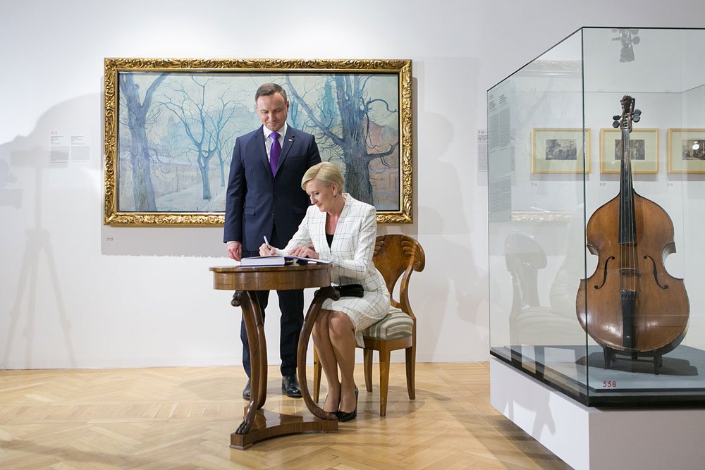 Kobieta o krótkich blond włosach w białym żakiecie i spódnicy siedzi przy małym okrągłym drewnianym stoliku i pisze w książce, obok stoi mężczyzna w ciemnym garniturze i fioletowym krawacie, za nimi na białej ścianie wisi obraz w złotej ramie przedstawiający nagie drzewa, a po prawej w szklanej gablocie eksponowany jest kontrabas, wszystko na drewnianym parkiecie galerii.