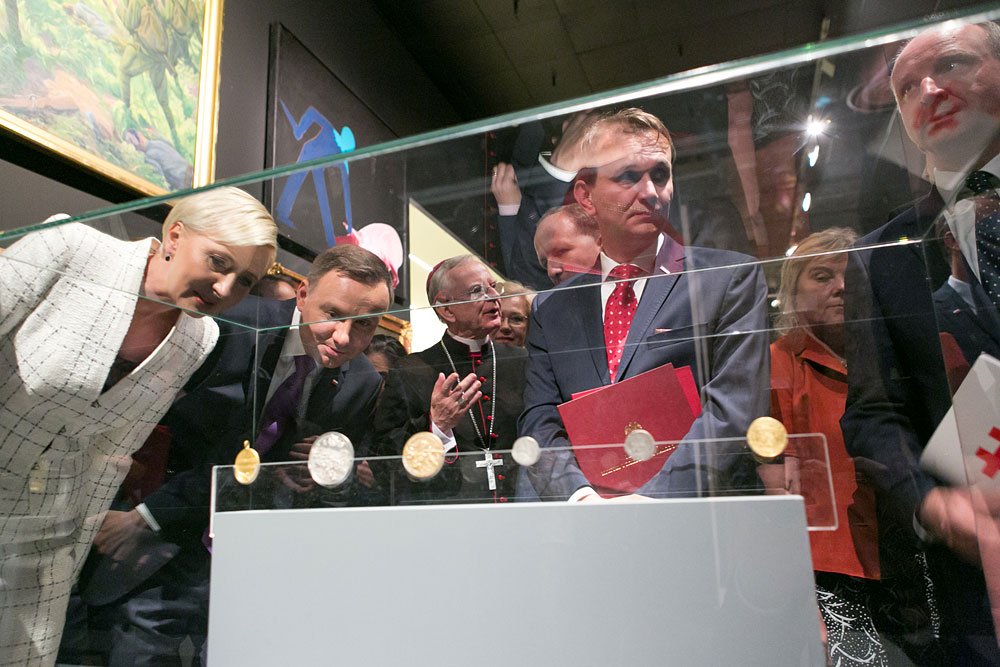 Grupa osób w eleganckich strojach pochylona nad szklaną gablotą z rzędem starych monet lub medali, w tle obrazy na ścianie i widoczny duchowny w sutannie z krzyżem.