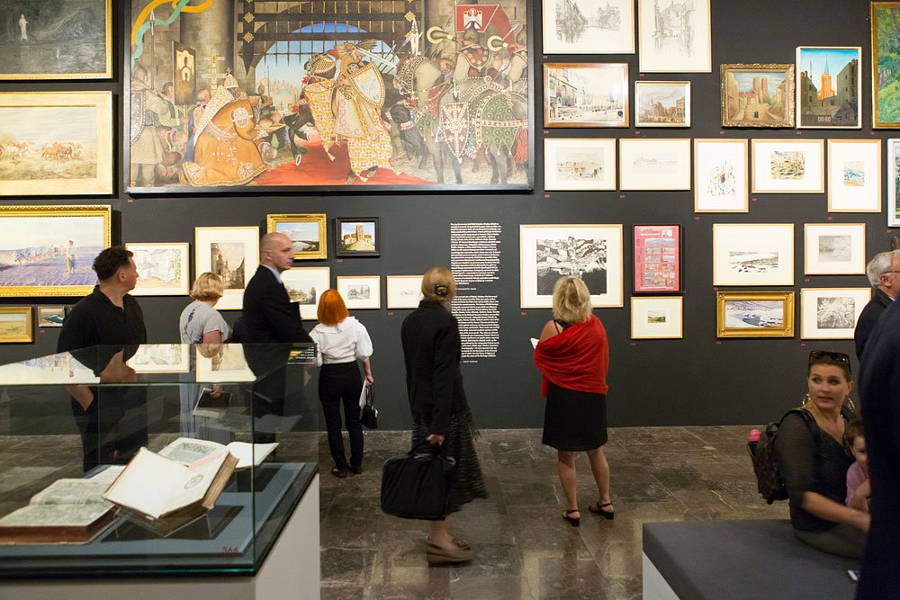 Wnętrze galerii sztuki z ciemną ścianą wypełnioną licznymi oprawionymi rysunkami i obrazami, dużym barwnym dziełem o motywie rycerskim u góry, szklaną gablotą z otwartą księgą na pierwszym planie oraz kilkoma osobami stojącymi i spacerującymi po kamiennej posadzce.
