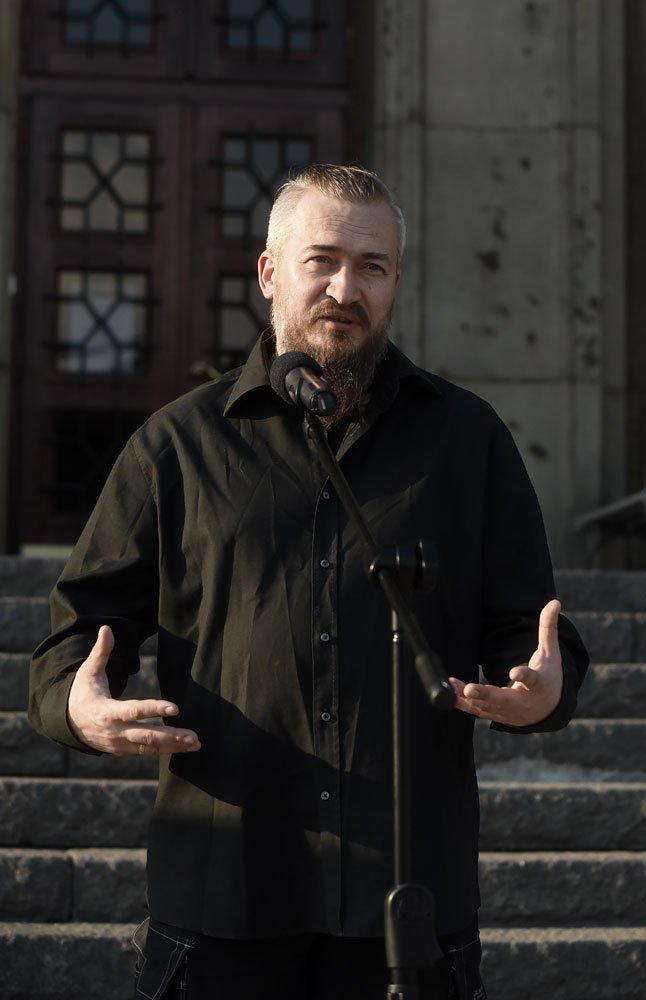 Mężczyzna z krótkimi jasnymi włosami i brodą w czarnej koszuli stoi na kamiennych schodach przed dużymi drewnianymi drzwiami i mówi do mikrofonu na statywie, gestykulując obiema rękami.