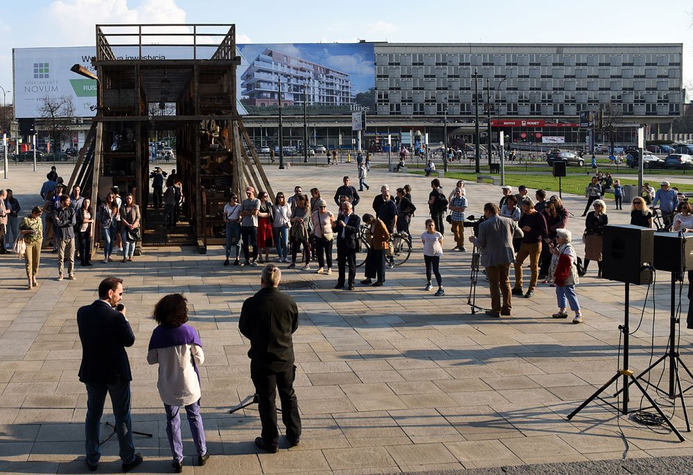 Widok na plac miejski: na pierwszym planie trzy osoby stojące przy mikrofonach i głośnikach na statywach, dalej grupa obserwujących i kilka osób z rowerami zgromadzonych przed dużą drewnianą, wielopoziomową instalacją, a w tle wielopiętrowy budynek z dużym billboardem reklamowym.