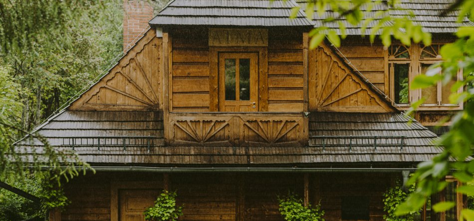 Fasada drewnianego domu w otoczeniu zieleni z gontowym dachem, centralną lukarną z wąskimi drzwiami i rzeźbionymi trójkątnymi ornamentami pod okapem, prawym oknem z małymi szybami, ceglastym kominem po lewej oraz pnączami i liśćmi na pierwszym planie, w padającym deszczu.