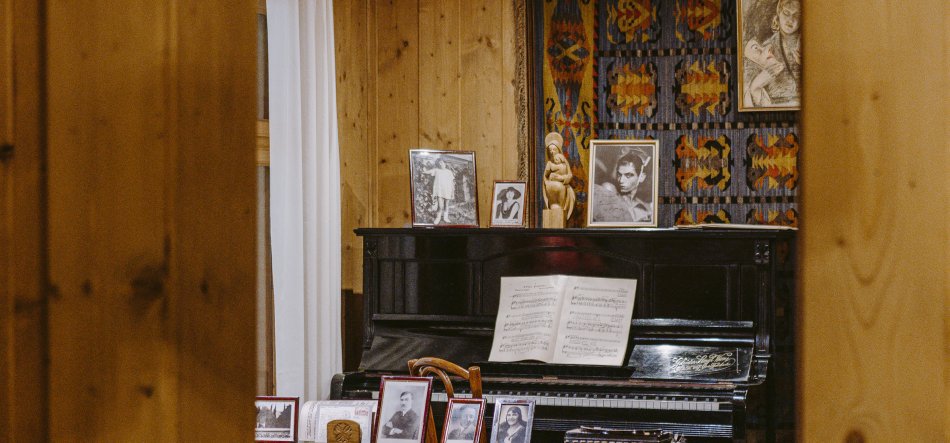 Wnętrze z drewnianymi panelami i widocznymi framugami po bokach; czarne pianino z otwartą nutą, na jego wieczku ustawione oprawione fotografie i drobna rzeźbiona figurka, a za nim kolorowy geometryczny kilim na ścianie i biała zasłona po lewej.