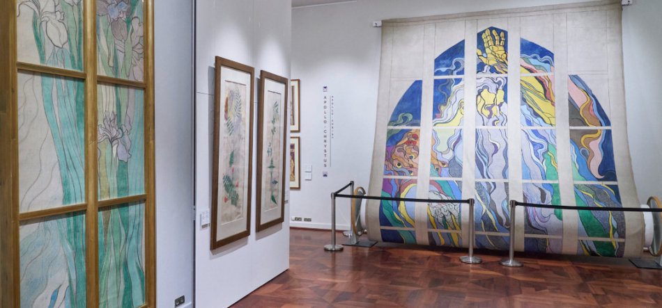 Wnętrze galerii z połyskującą drewnianą podłogą; po lewej pionowy panel w drewnianej ramie ze szkicami zielono‑niebieskich kwiatów, na centralnej białej ścianie trzy podłużne oprawione rysunki botaniczne, a po prawej duża wisząca wielopanelowa praca o abstrakcyjnych, falistych wzorach w żywych pastelowych i intensywnych kolorach odgrodzona niskimi słupkami z linami.