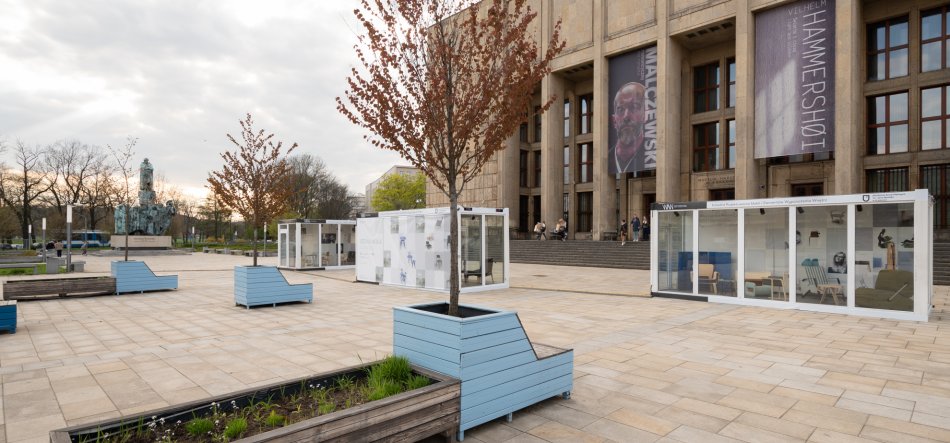 Rozległy kamienny plac przed monumentalnym budynkiem z wysokimi oknami i pionowymi banerami, na którym rozmieszczone są przezroczyste szklane pawilony wystawowe, niebieskie drewniane ławki z wbudowanymi donicami i młodymi drzewami, po lewej pomnik i w tle kilka osób stojących na schodach wejścia.