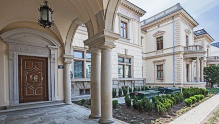 Fasada budynku z arkadowym podcieniem i kolumnami, ozdobnymi drewnianymi drzwiami po lewej, wiszącą latarnią, szeregiem okien oraz niewielkim zadbanym ogrodem i alejką z prawej.