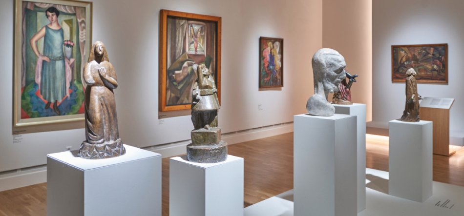 Galeria muzealna z białymi podestami prezentującymi kilka rzeźb o różnych formach — stojące figury, wielostopniowa abstrakcyjna forma i szare popiersie — oraz z kilkoma obrazami w ramach na jasnych ścianach, drewnianą podłogą i punktowym oświetleniem.