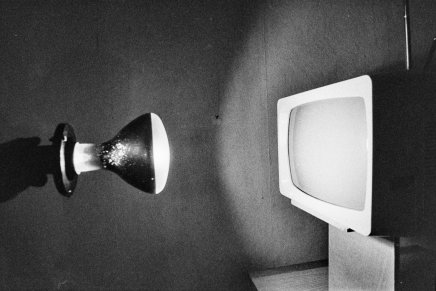 Czarno-białe zdjęcie z lewostronnie zamocowaną lampą o szerokim kloszu rzucającą światło i cień na ścianę oraz po prawej starym telewizorem kineskopowym o jasnej obudowie ustawionym na niewielkim meblu.