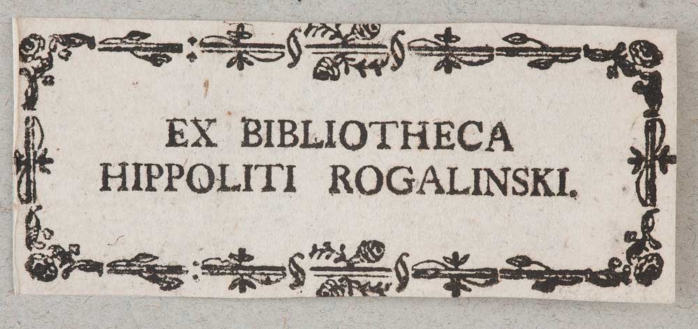 Prostokątna biała papierowa etykieta z czarnym wersalikowym napisem EX BIBLIOTHECA HIPPOLITI ROGALINSKI wyśrodkowanym i otoczonym czarną ornamentalną ramką z kwiatowo‑liściastymi elementami oraz drobnymi przebarwieniami papieru na szarym tle.