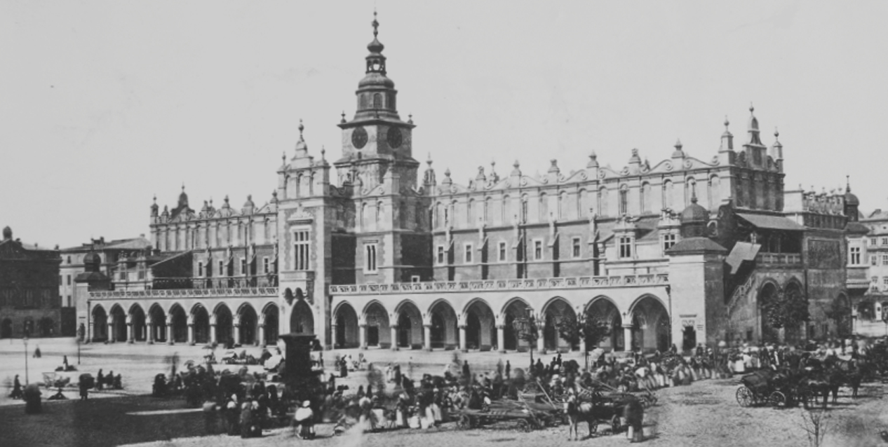 Czarno-białe zdjęcie rozległego zabytkowego gmachu z rzędem arkad na parterze, wysoką centralną wieżą z tarczami zegara i dekoracyjnymi szczytami, stojącego przy dużym rynku, na którym widać tłum ludzi i zaprzęgi konne.