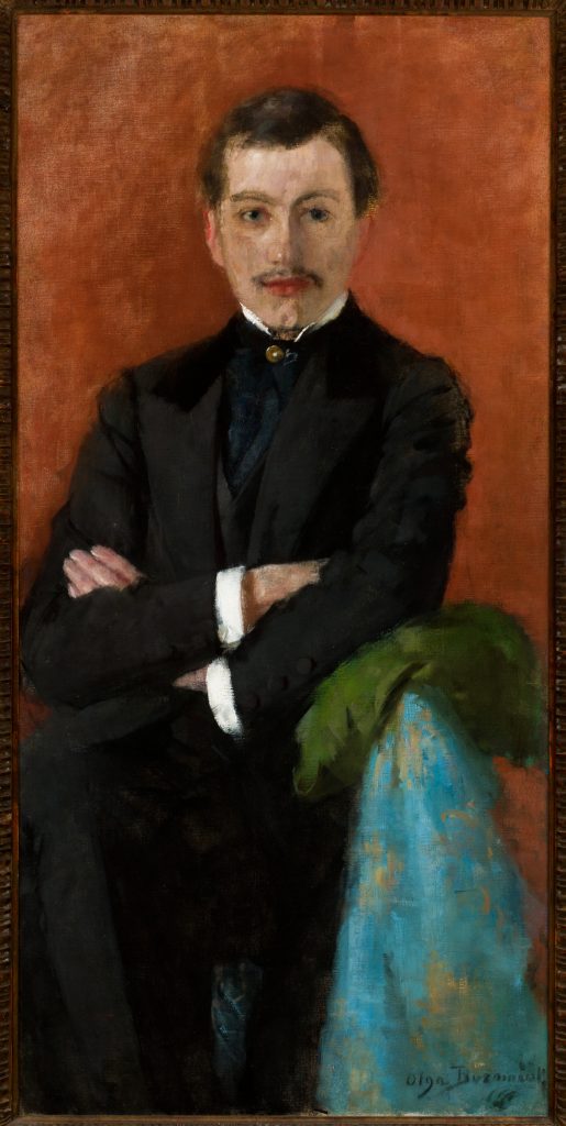 Portret olejny przedstawiający mężczyznę o krótkich ciemnych włosach i wąsach, siedzącego z założonymi rękami w czarnym garniturze na jednolitym ciepłym czerwono-brązowym tle, z zielonym i niebieskim materiałem na poręczy oraz wąską ramą obrazu widoczną na krawędziach.