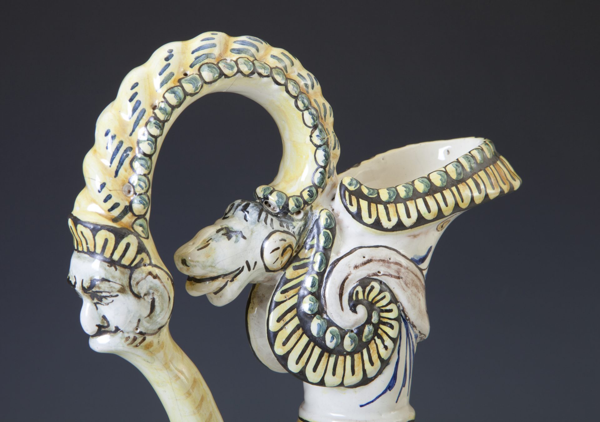 Zbliżenie fragmentu ozdobnego ceramicznego naczynia z profilową twarzą na uchwycie i rzeźbioną głową zwierzęcą z spiralnym rogiem, kremowe szkliwo ozdobione żółto‑zielono‑czarnymi malowanymi wzorami i ornamentami na ciemnym tle.