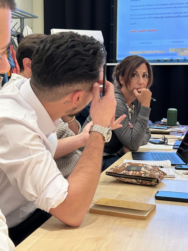 Grupa dorosłych siedzących przy stole z notatnikami i laptopem; zbliżenie na mężczyznę w białej koszuli widzianego z boku trzymającego długopis przy głowie oraz kobietę naprzeciwko z ręką przy ustach i długopisem, na stole kwiatowa kosmetyczka, notesy, telefon i zielony bidon, w tle ekran projekcyjny i wieszak na ubrania.