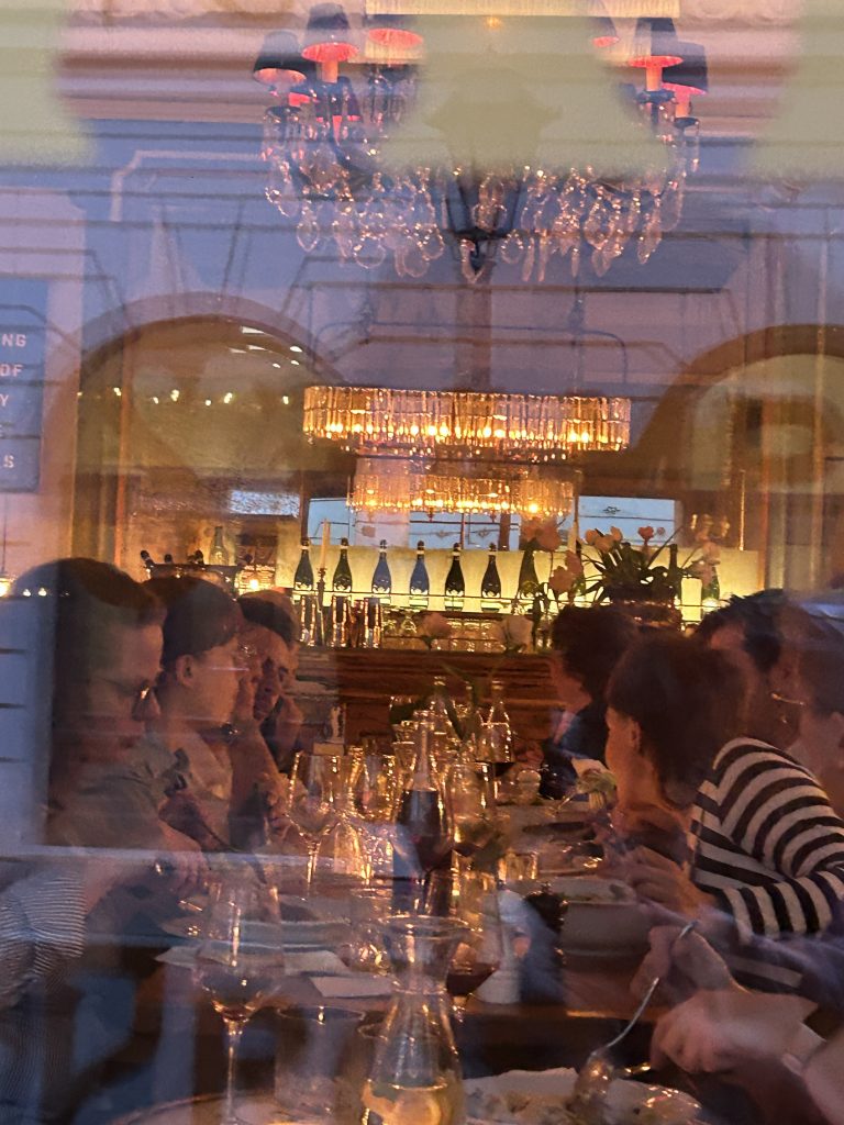 Odbicie przez szybę pokazuje wnętrze restauracji z kryształowym żyrandolem u góry i prostokątnym kryształowym żyrandolem nad barem, rzędem butelek na półce, licznymi błyszczącymi kieliszkami na stole oraz zarysami kilku osób siedzących przy wspólnym stole, częściowo nakładające się warstwy odbić i ciepłe pomarańczowe światło.