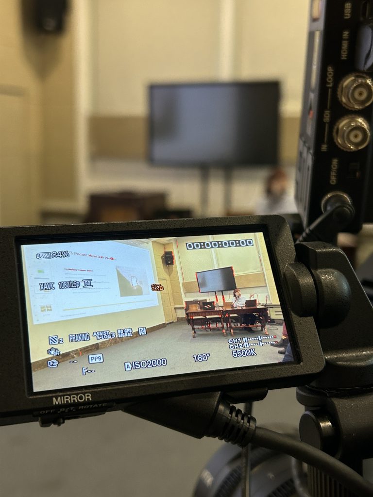 Zbliżenie na ekran podglądu kamery pokazujący wnętrze sali konferencyjnej: po lewej projekcja slajdów, w środku telewizor na stojaku i osoba siedząca przy stole, po prawej fragment korpusu kamery ze złączami, a na ekranie widoczne ikony ustawień kamery (bateria 84%, ISO2000, 5500K, licznik czasu).