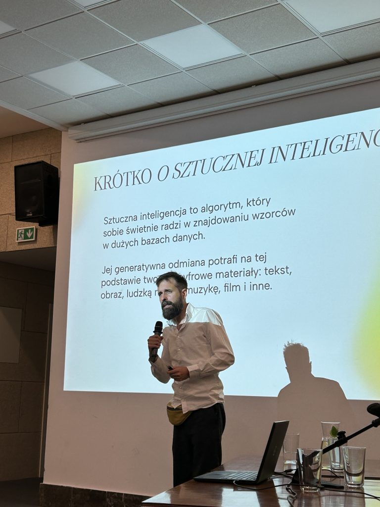 Mężczyzna z mikrofonem w białej koszuli stoi przed dużym ekranem projekcyjnym wyświetlającym slajd z nagłówkiem KRÓTKO O SZTUCZNEJ INTELIGENCJI i polskim tekstem, jego cień pada na ekran; na pierwszym planie stół z otwartym laptopem, szklankami i mikrofonem, a w tle sufit z paneli akustycznych i głośnik ścienny.