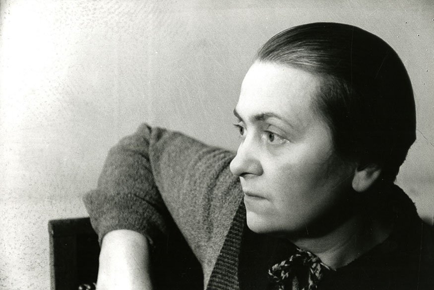 Czarno-białe zdjęcie w profilu przedstawiające kobietę ze ściśle zaczesanymi włosami, w swetrze i apaszce związanej pod szyją, z ramieniem opartym o oparcie krzesła na gładkim, jasnym tle.