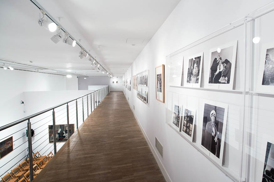 Długi, jasny korytarz antresoli galerii z drewnianą podłogą i metalową balustradą po lewej, białą ścianą po prawej z oprawionymi czarno-białymi fotografiami, sufitowymi reflektorami oświetlającymi ekspozycję oraz widokiem na niższą kondygnację z krzesłami i obrazami.