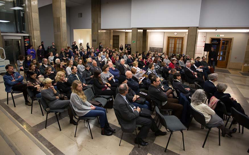 Duża grupa mężczyzn i kobiet w różnym wieku siedzących w rzędach czarnych krzeseł w jasnej, marmurowej sali z wysokimi kolumnami i podwójnymi drzwiami w tle, kilka osób stoi przy ścianie.