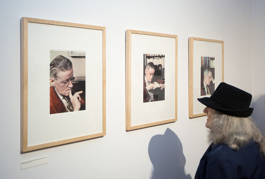 Trzy pionowe fotografie w jasnych drewnianych ramach z białymi paspartu wiszą równo na białej ścianie galerii, każda ukazuje starszego mężczyznę w okularach w różnych pozach, po prawej stoi zwrócona ku nim osoba w czarnym kapeluszu i ciemnym płaszczu oraz jej cień na ścianie, pod lewą ramą widoczna mała tabliczka.