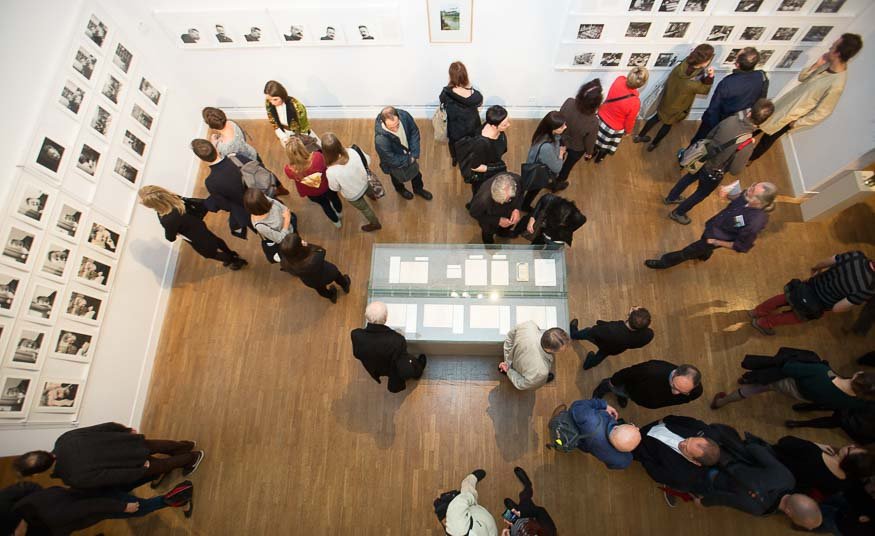 Zdjęcie z lotu ptaka wnętrza galerii: kilkadziesiąt osób ogląda oprawione fotografie rozmieszczone w siatce na białych ścianach, a pośrodku na drewnianej podłodze stoi przeszklona gablotka z ułożonymi kartkami i materiałami wystawowymi.