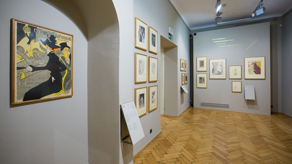 Wnętrze jasnej galerii z łukowym przejściem po lewej, szarymi ścianami i jodełkową drewnianą podłogą, na których wiszą liczne oprawione rysunki i plakaty (po lewej duży żółto-czarny plakat), przy ścianach ustawione tablice informacyjne, a sufitowe reflektory i rzutnik oświetlają ekspozycję i wyświetlają tekst na tylnej ścianie.