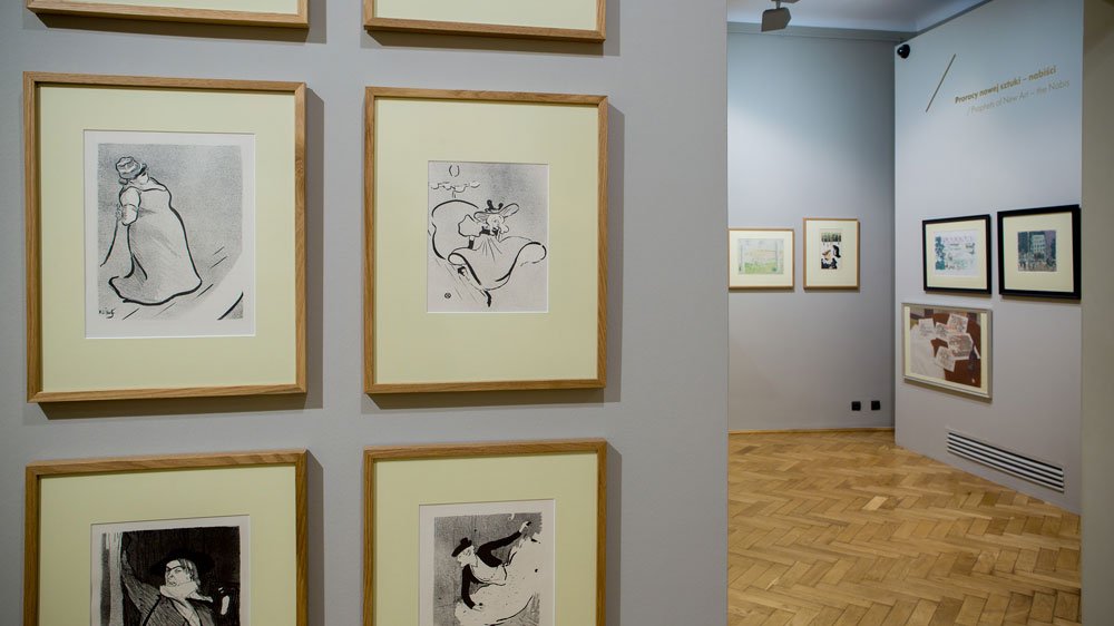 Wnętrze galerii z szarymi ścianami i drewnianą mozaikową podłogą; z przodu po lewej widoczne kilka oprawionych czarno-białych rysunków w jasnych passe-partout i prostych drewnianych ramach, a w głębi po prawej przejście do kolejnej sali z dodatkowymi obrazami powieszonymi na ścianie i niewyraźnym napisem.